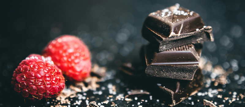 Três quadrados de chocolate negro uns em cima dos outros ao lado de duas framboesas vermelhas num fundo preto com crostas de chocolate e açúcar branco.