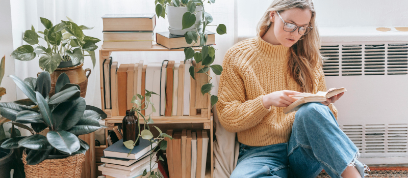 rapariga sentada no chão a ler e ao lado de uma estante de livros com plantas